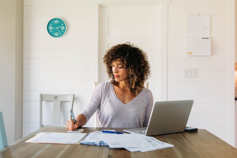 Woman writing notes at a desk with paperwork and a laptop Woman doing finances at home